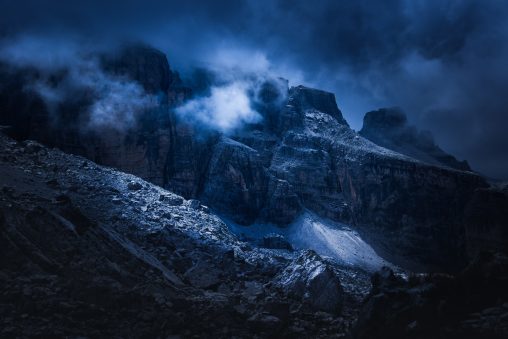 Ghiaione illuminato in primo piano e grandi pareti rocciose tra le nubi, fotografia di paesaggio a toni freddi per il progetto The Dark Side.